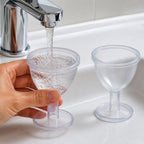 A hand holds a clear eye wash cup under a running faucet, filling it with water, while another empty cup sits on the white