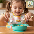 A young girl in a bib holds a panda-handled fork and spoon, eating pasta from a teal bowl.