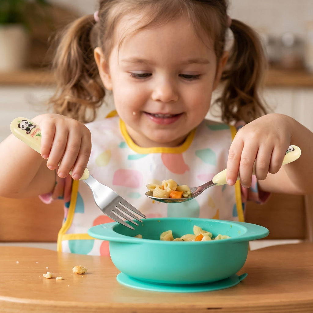 A young child with pigtails uses a yellow panda spoon and fork to eat pasta from a teal silicone bowl.