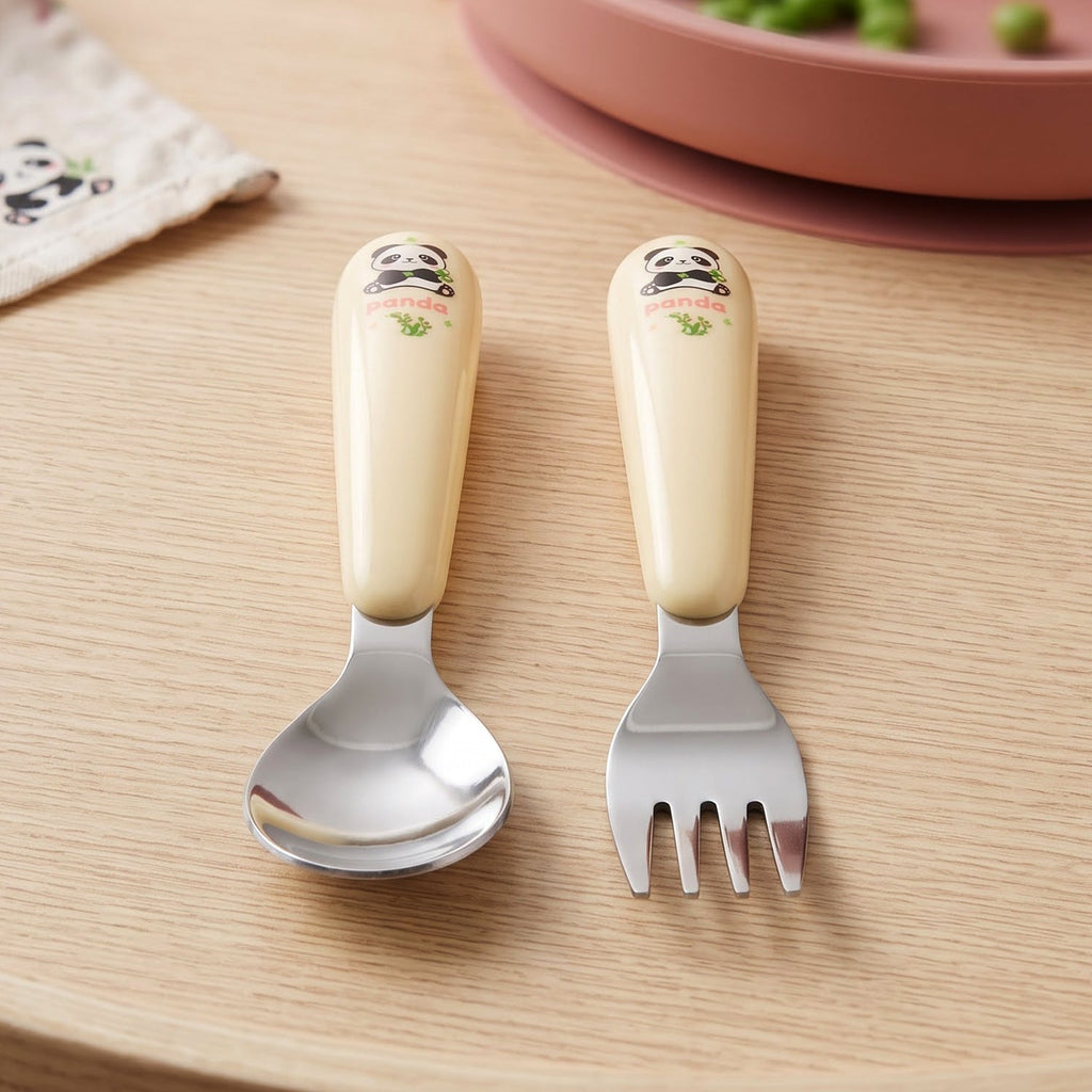 A yellow and silver panda-themed spoon and fork set rests on a wooden surface beside a pink plate.