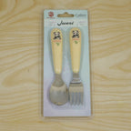 A yellow and silver panda-themed spoon and fork set is sealed in clear plastic packaging on a light wooden surface.
