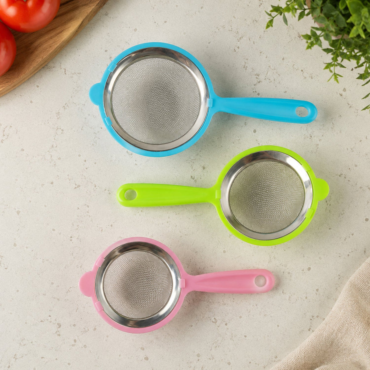Three individual strainers with blue, green, and pink plastic handles and stainless steel mesh lie separately on a