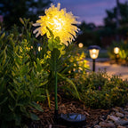 A white chrysanthemum flower solar stake light glowing in a garden at twilight, with other garden lights visible in the