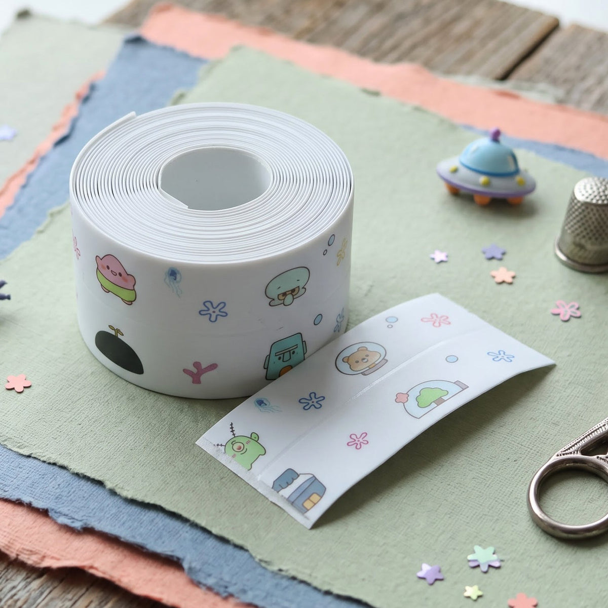 A roll of white cartoon-printed sealing tape and a cut piece of tape on textured paper with craft supplies.