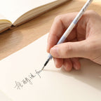 A hand writes in a foreign language on a light-colored paper with a white and blue pen refill, with a notebook.