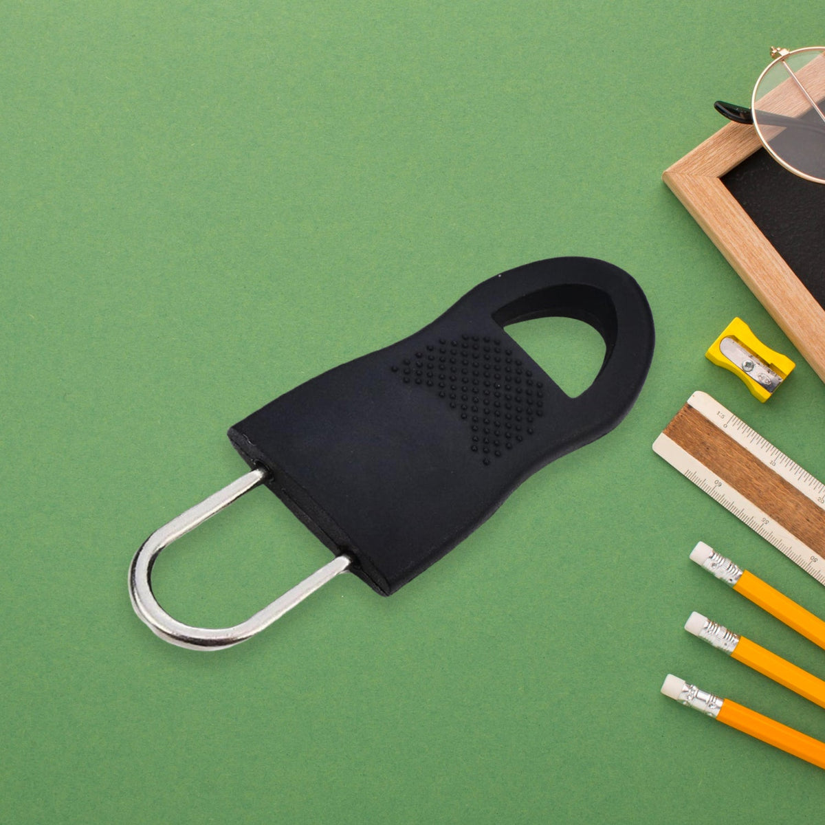 A single black rubber zipper pull tab on a green background with school supplies like pencils and a ruler.