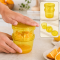 A person's hands press down on a yellow plastic manual food grinder filled with orange segments, extracting juice. The inset