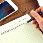 A hand holding a black 3-in-1 stylus pen, writing in a notebook next to a smartphone.