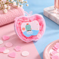 A clear, apple-shaped container filled with small pink and white paper soap pieces, on a pink and blue surface with a towel
