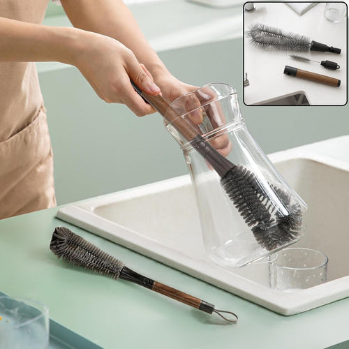 A person cleans a clear glass carafe with a long-handled grey brush, while a second brush rests on the counter.