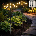 Two solar-powered firefly garden stake lights illuminate a lush green garden path at dusk.