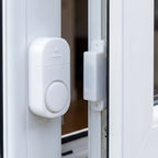 A white plastic magnetic alarm is attached to a white window frame, with its smaller magnetic component on the adjacent