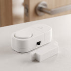 The white magnetic door alarm and its smaller magnetic piece are on a white surface, with a blurred wooden door in the