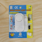A white magnetic door/window entry alarm is sealed in its blue and yellow retail packaging on a light wooden surface.
