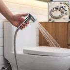 A hand holds a chrome bidet sprayer, spraying water towards a white toilet in a tiled bathroom.