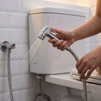 A hand holds a chrome bidet sprayer, spraying water next to a white toilet with a connected hose.