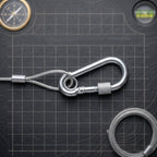 A close-up shows a silver carabiner hook attached to a braided stainless steel wire rope on a dark grid surface.