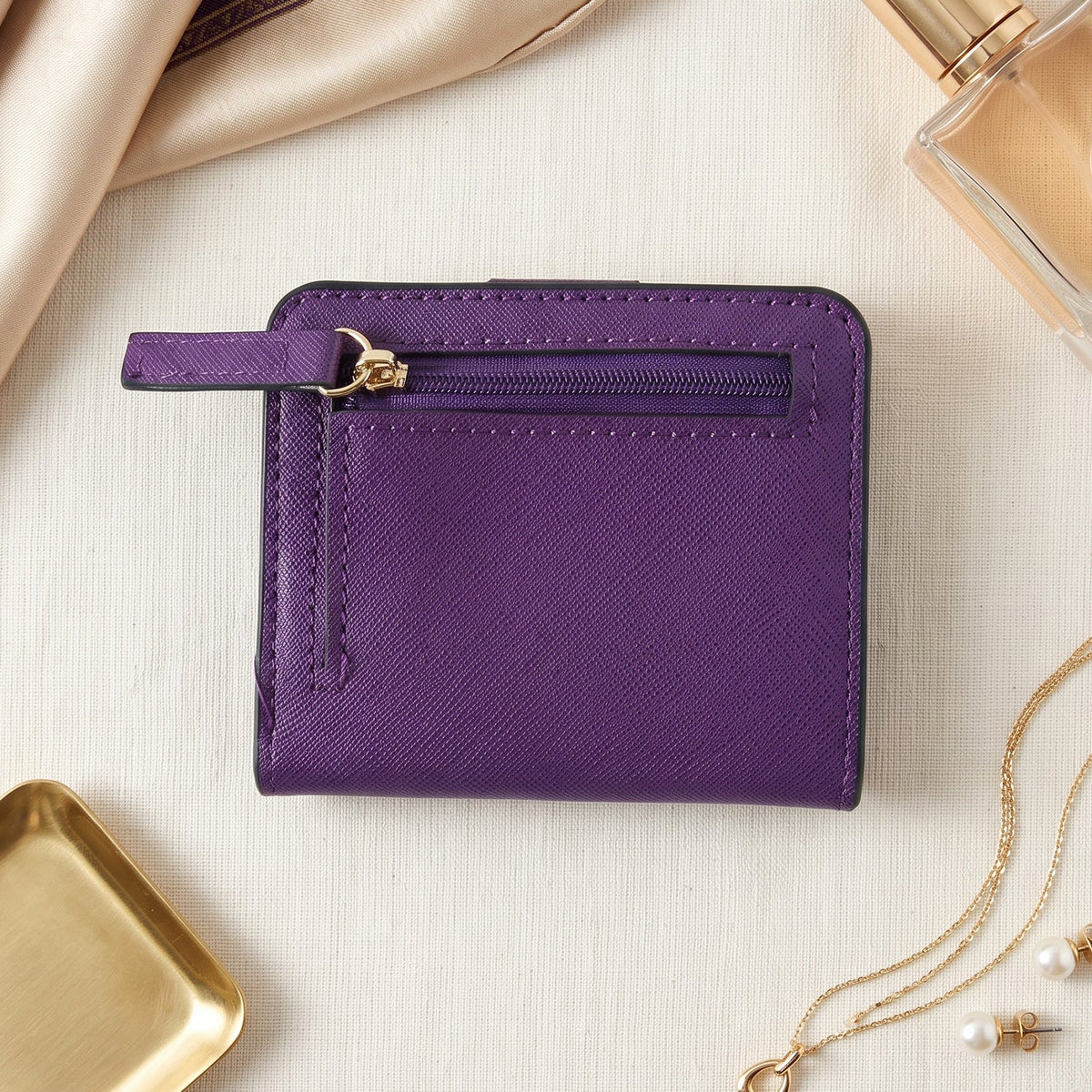 A closed purple bifold wallet with a gold-zippered coin pouch on the back, surrounded by jewelry and a perfume bottle.