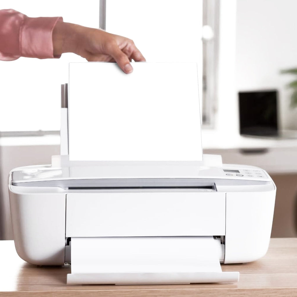 A person's hands feed a sheet of white paper into a white printer on a wooden desk.
