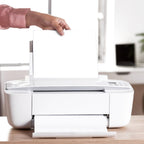 A person's hands feed a sheet of white paper into a white printer on a wooden desk.
