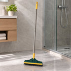 A green and yellow floor cleaning brush with a metal handle stands on a grey tiled bathroom floor, next to a shower and