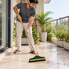 A man in a grey shirt and beige pants sweeps dirt on a tiled balcony with a green and yellow brush, surrounded by potted