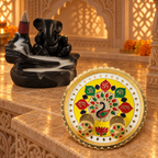 A black Ganesha backflow incense burner with smoke and a yellow and gold puja thali with peacock design on a marble surface.