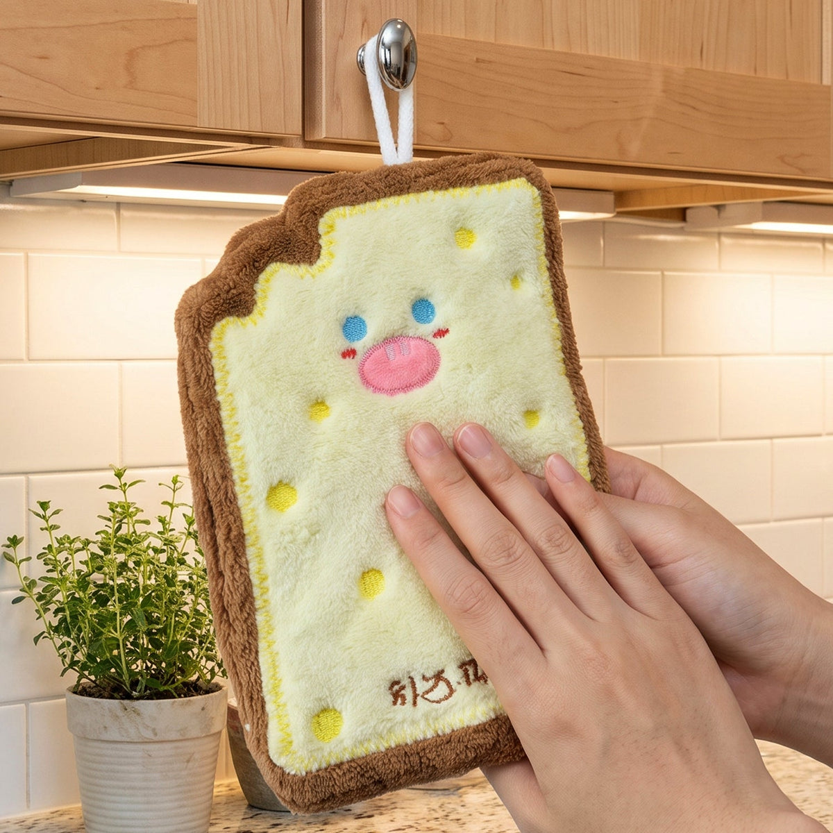 A person's hands are shown drying on a cartoon cheese toast-shaped microfiber towel hanging from a kitchen cabinet knob.