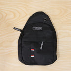 A black sling bag with a front pocket and 'FASHION INSTINCT' label, lying flat on a light wooden surface.
