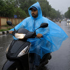 A man wearing a blue rain poncho and a baseball cap rides a black scooter on a wet, rainy street.