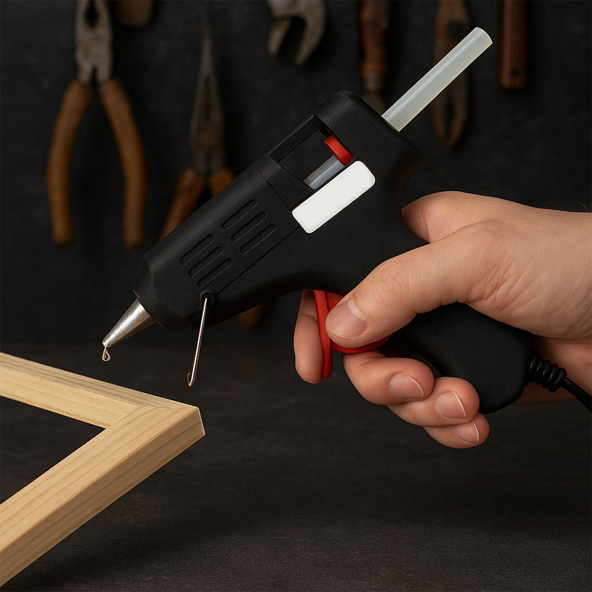 A hand holding a black and red glue gun, with a drop of hot glue dripping from the nozzle onto a wooden frame.
