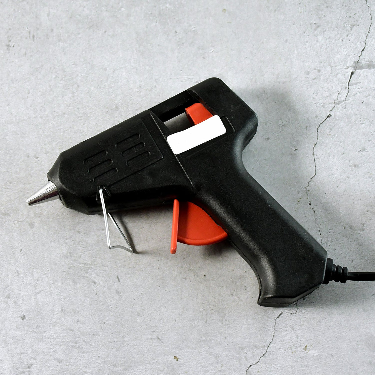 A black and red glue gun with a white label and a metal stand, resting on a gray concrete surface.