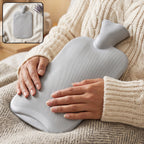 A person in a cream sweater holds a grey ribbed hot water bottle on their lap, with a small inset showing the bottle alone.