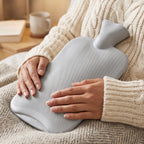 A person in a cream sweater holds a grey ribbed hot water bottle on their lap, with a blurred background of a mug and book.