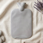 A grey ribbed hot water bottle lies flat on a light fabric, with lavender sprigs and smooth white stones beside it.
