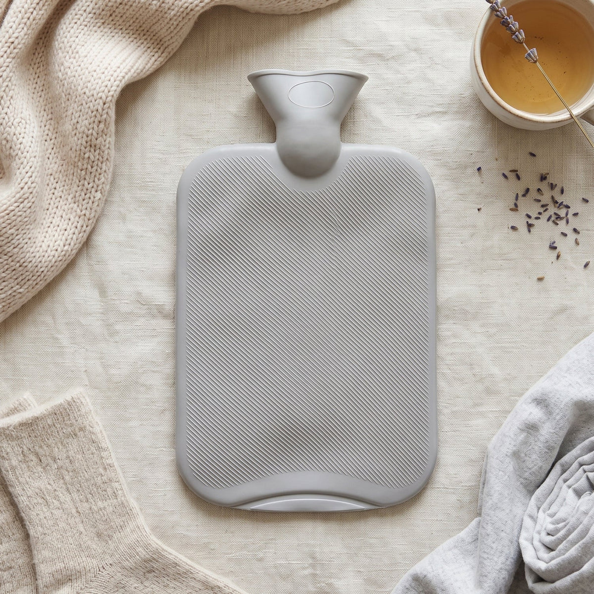 A grey ribbed hot water bottle is centered on a light fabric, surrounded by a knitted scarf, socks, folded fabric, and a cup