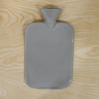 A grey ribbed hot water bottle with a textured surface and wide neck is placed on a light wooden background.