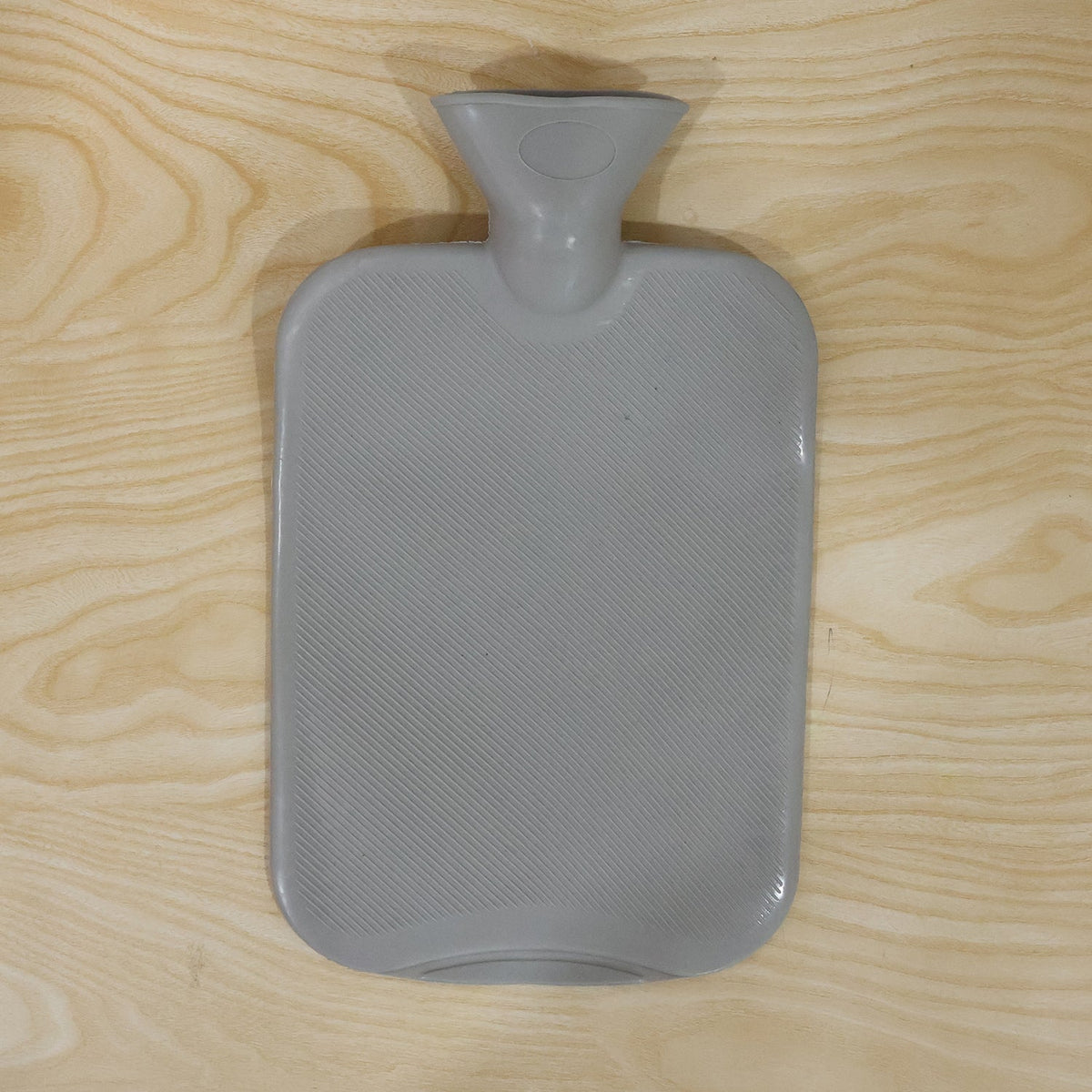 A grey ribbed hot water bottle with a textured surface and wide neck is placed on a light wooden background.