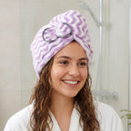 A smiling woman with wet, curly hair and a purple and white chevron hair towel wrap on her head in a bathroom.