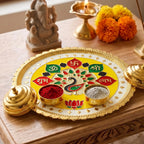 A yellow and gold puja thali with peacock design, small bowls of red and grey powder, and a Ganesha statue in the background.