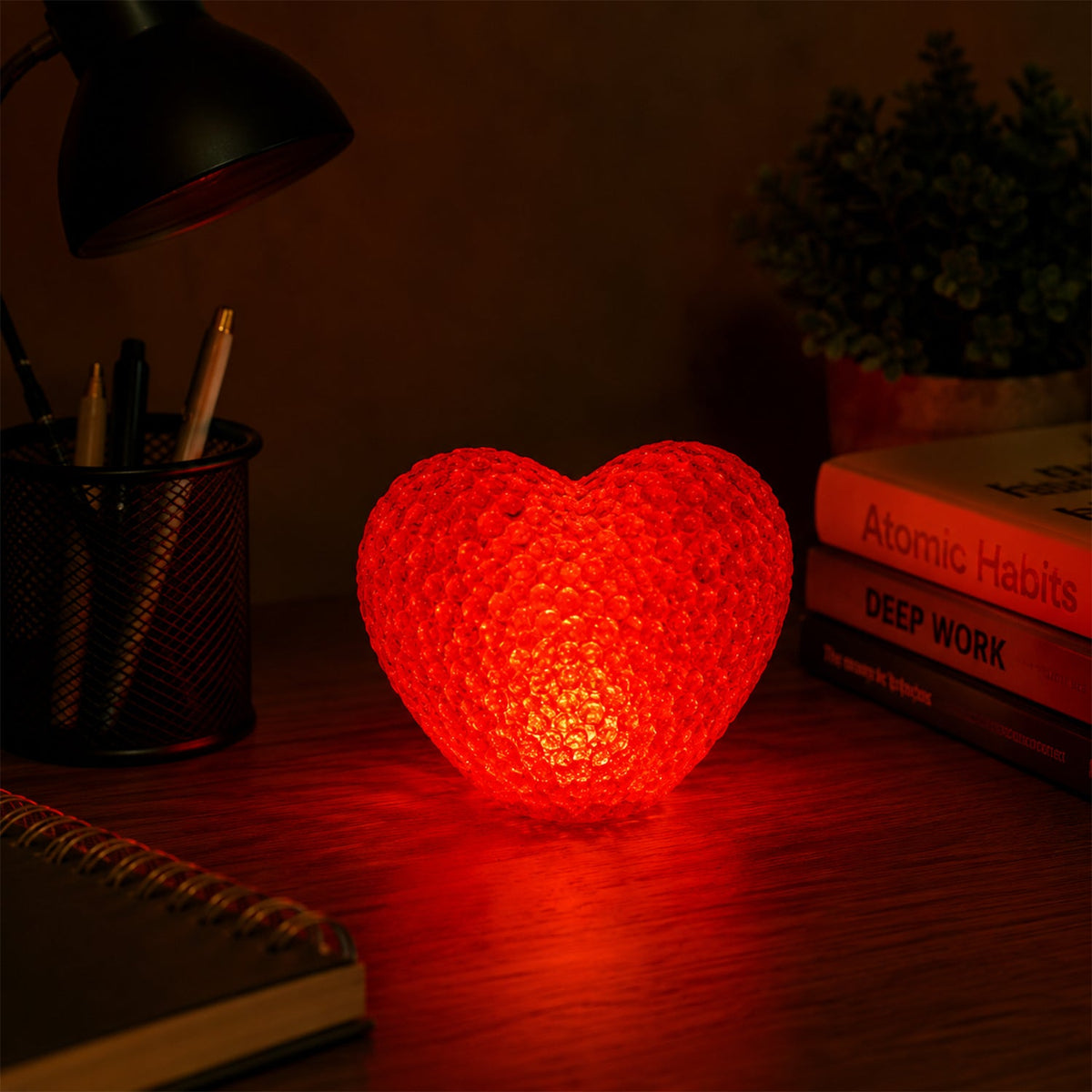 A red heart-shaped LED night lamp casts a soft glow on a dark desk beside a black lamp and stacked books.