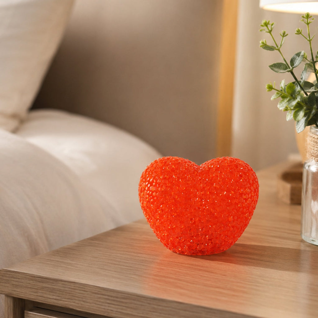 A red heart-shaped LED night lamp rests on a wooden nightstand beside a small plant in a clear vase.