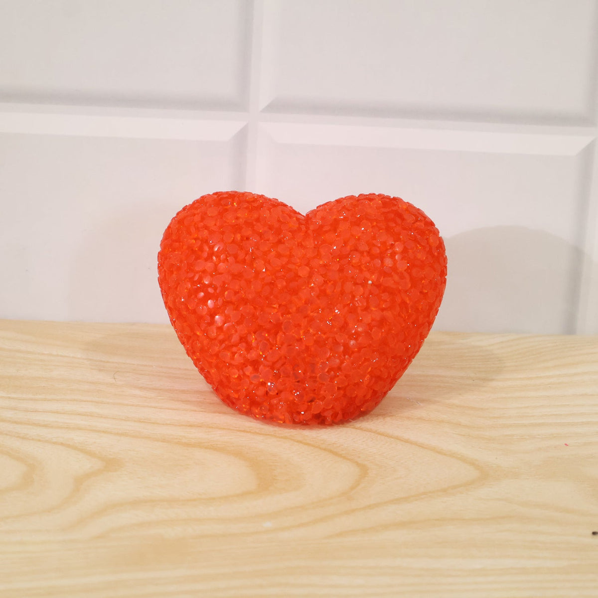 A textured orange heart-shaped LED lamp is centered on a light wooden surface against a white tiled wall.