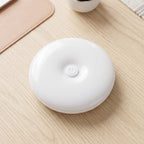 A white, round LED light with a central power button rests on a light wood desk next to a pink mat