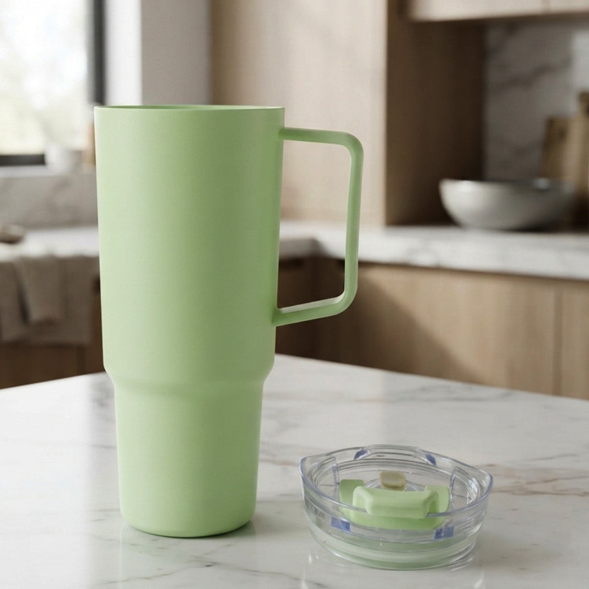 A light green travel tumbler with a handle stands on a marble counter next to its clear lid in a kitchen.