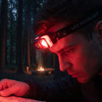 A man wearing an orange and black headlamp with a red light, reading a map in a dark forest with a campfire in the – view 7