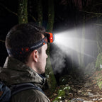 A man from behind wearing an orange and black headlamp with a bright white beam illuminating a dark forest path.
