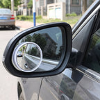 A grey car's side mirror with a small round blind spot mirror attached, reflecting a street scene.