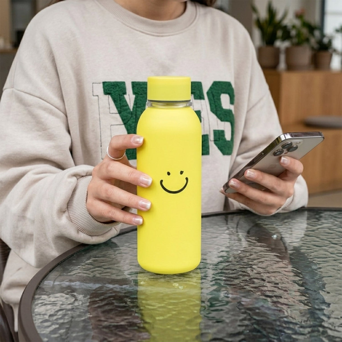 A person holds a yellow water bottle with a smiley face and a smartphone at a glass table.