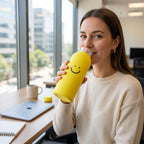 A woman smiling and drinking from a yellow glass water bottle with a smiley face at an office desk.